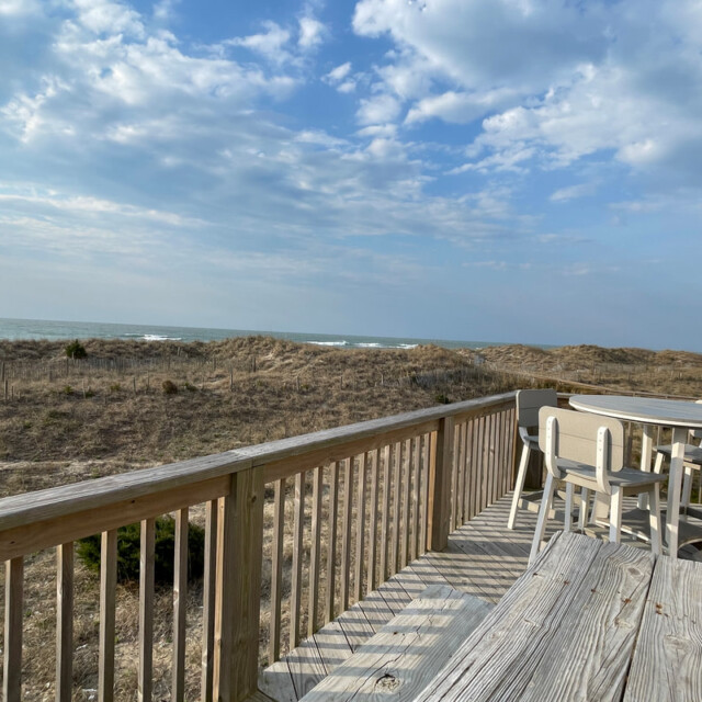 Wooden deck with round table and chairs overlooks sandy dunes and ocean under a partly cloudy sky.