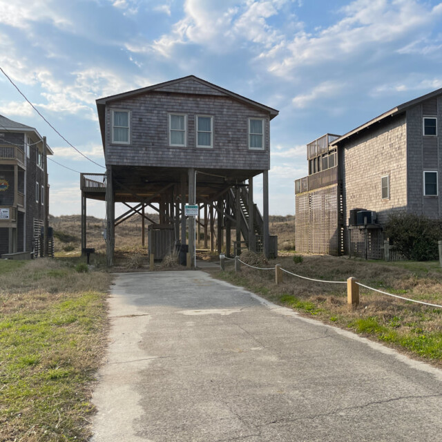 Wooden beach house on stilts at the end of a concrete driveway, with grassy yard and partly cloudy sky.