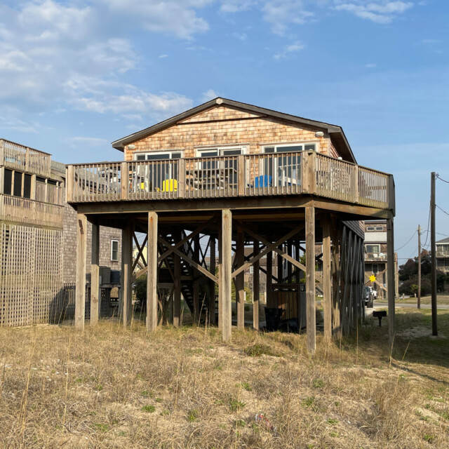 Elevated beach house with wooden stilts, deck, and outdoor chairs, surrounded by dry grass and neighboring houses.
