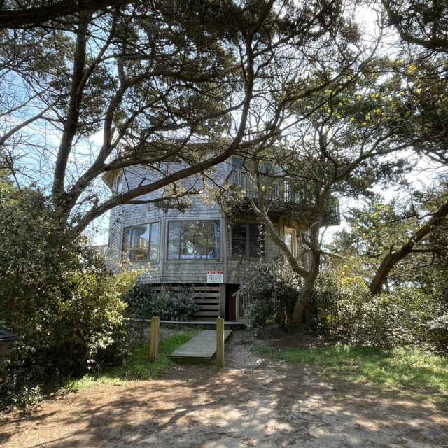 A two-story wooden house surrounded by trees, with a ramp leading to the front door and a visible “for rent” sign.