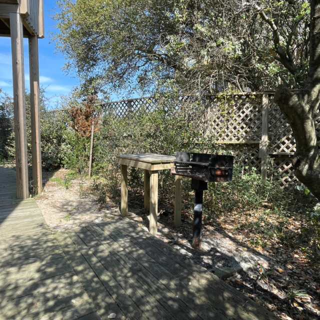 Wooden walkway beside an outdoor grill, table, and trees, with a lattice fence in the background on a sunny day.