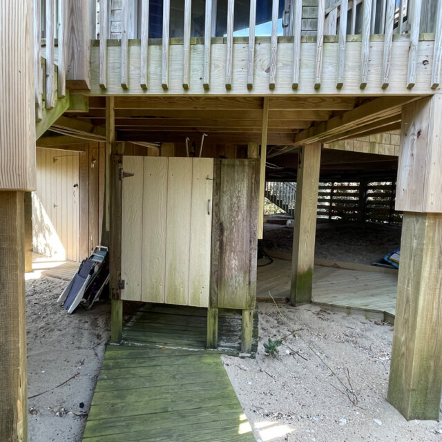 Wooden outdoor shower under a raised deck with sand, a boardwalk, and some beach equipment nearby.