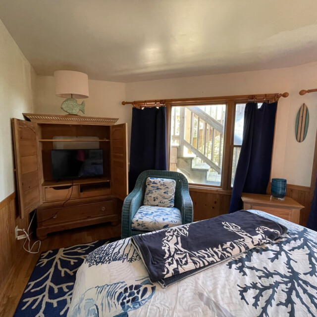 Cozy bedroom with a bed, TV in wooden cabinet, blue chair, and window showing wooden stairs outside.