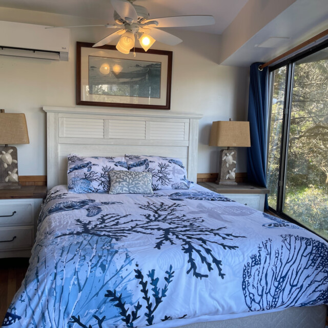 A neatly made bed with sea-themed bedding, two lamps, and large windows overlooking trees in a bright bedroom.