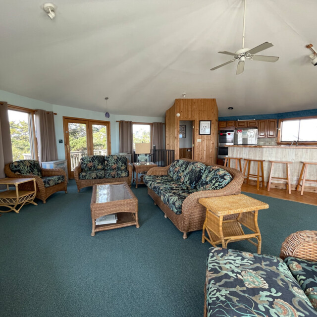 Spacious living room with wicker furniture, green carpet, and open kitchen with bar stools and wooden cabinets.
