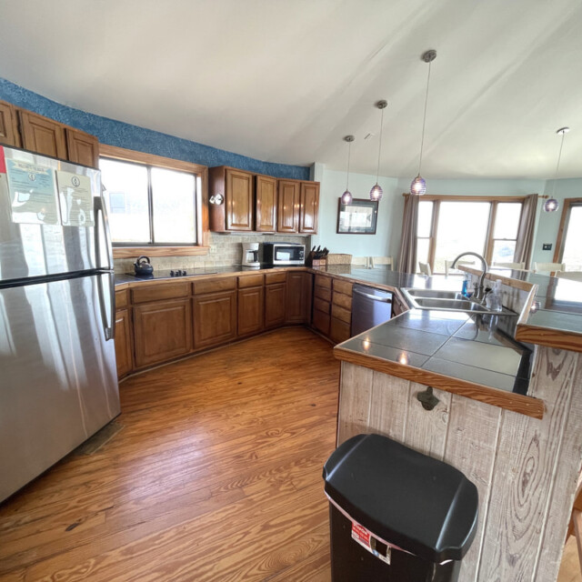 Spacious kitchen with wood cabinets, stainless steel fridge, bar seating, pendant lights, and large windows.