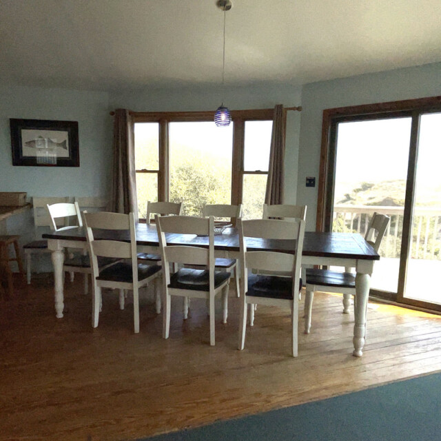 Dining room with a long table, eight chairs, wood floor, large windows, and sliding glass door to a deck.