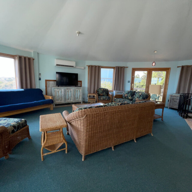 Spacious living room with wicker furniture, blue carpet, large windows, TV, and sliding glass doors to a deck.