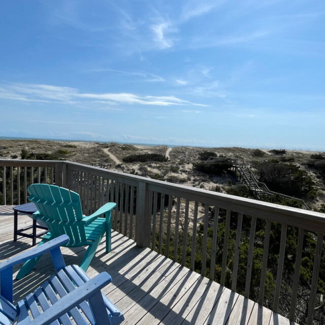 Wooden deck with colorful chairs overlooks sand dunes and a boardwalk leading toward the beach under a blue sky.
