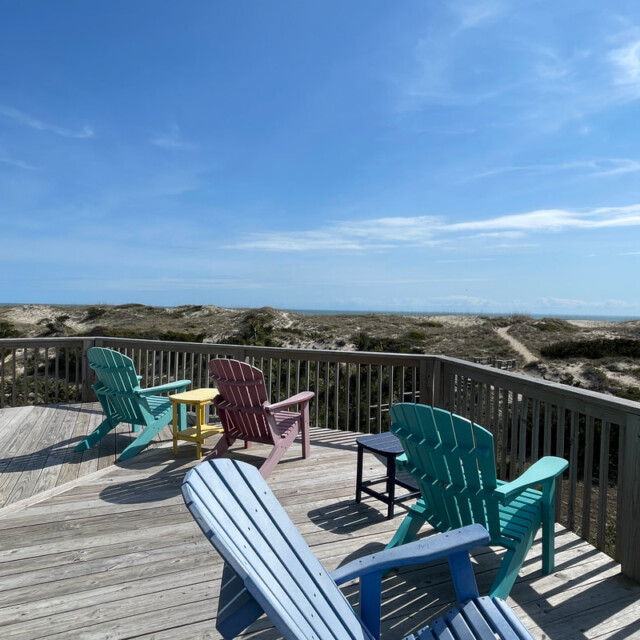 Five colorful Adirondack chairs on a wooden deck overlook sand dunes and blue sky at the beach.