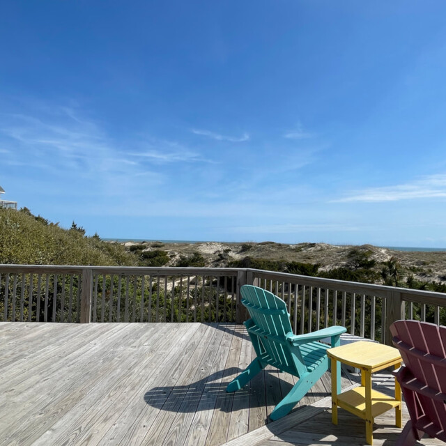 Wooden deck with colorful chairs and a small table overlooks sand dunes and a clear blue sky.