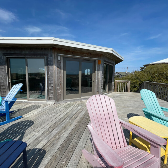 Wooden deck with colorful Adirondack chairs and a weathered house under a clear blue sky.