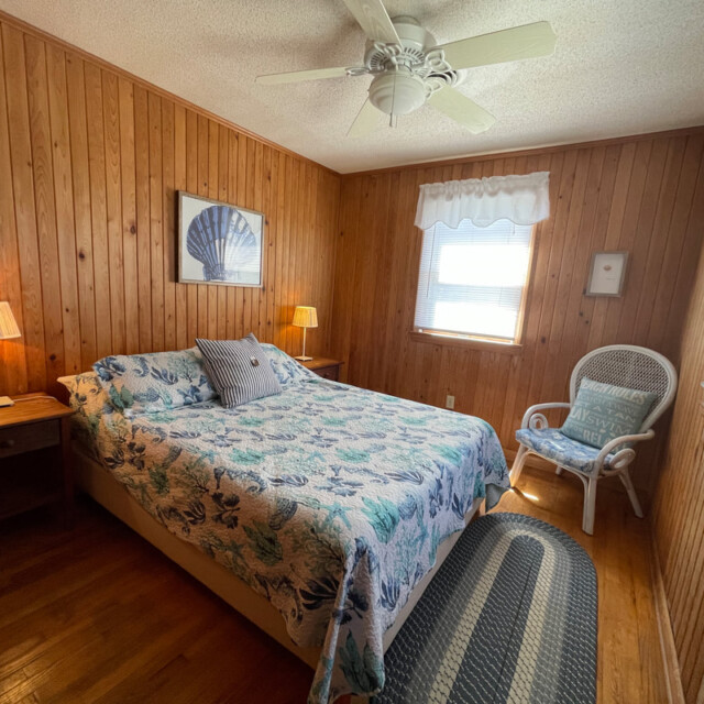 Wood-paneled bedroom with queen bed, wicker chair, two lamps, a ceiling fan, and light from two windows.