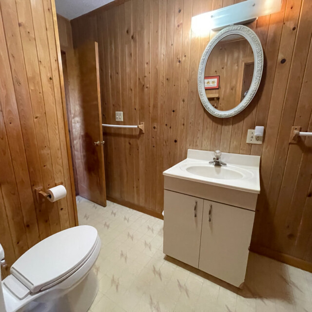 Bathroom with wood panel walls, white sink vanity, oval mirror, toilet, and open door.