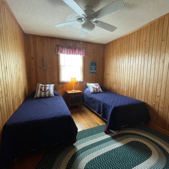 Wood-paneled bedroom with two twin beds, a window, a ceiling fan, and nautical-themed decor.