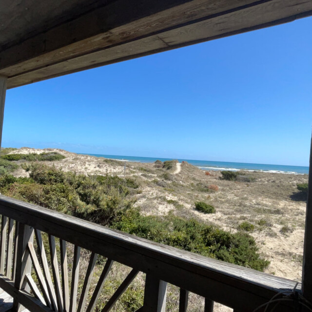View from a wooden porch overlooking sand dunes, green shrubs, and the ocean under a clear blue sky.