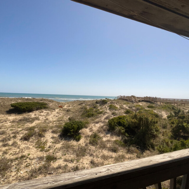 View of a sandy beach and ocean from a shaded wooden deck, with clear blue sky and some greenery visible.