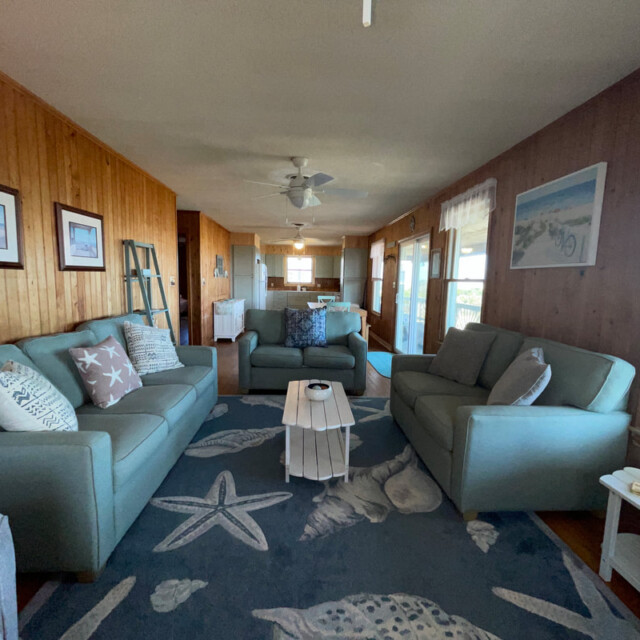 Living room with blue sofas, ocean-themed rug, wooden walls, framed art, and natural light from large windows.
