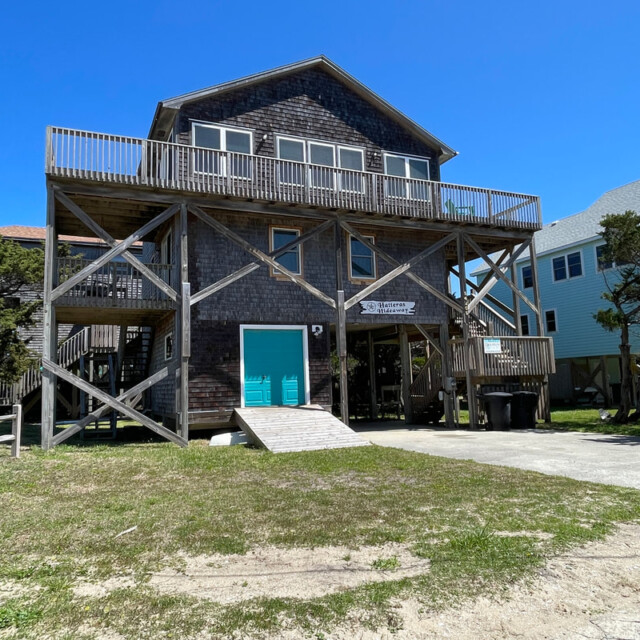 A two-story wooden beach house on stilts with a wraparound deck, driveway, and front steps under a clear blue sky.