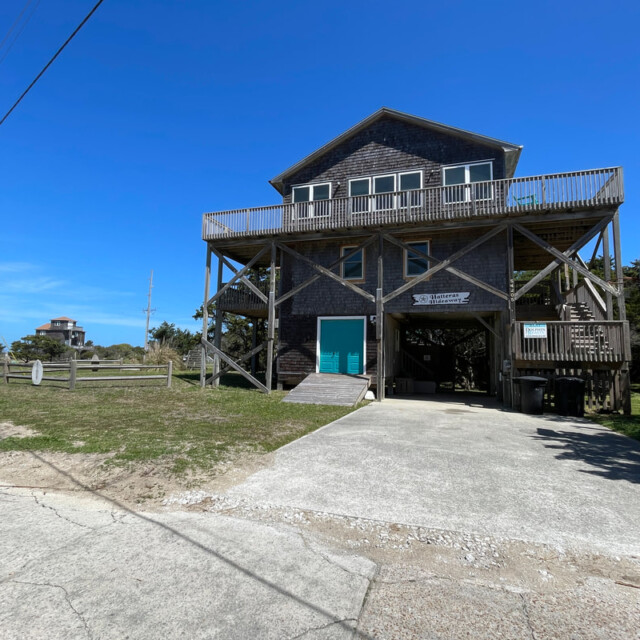 Two-story wooden beach house on stilts with driveway, upper deck, and clear blue sky in the background.