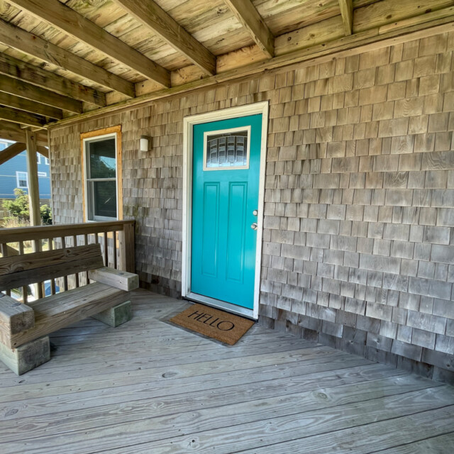 A turquoise door with a “Hello” doormat and a wooden bench on a covered porch with shingle siding.