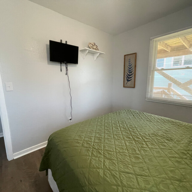 Small bedroom with a green bedspread, wall-mounted TV, window with blinds, and simple decor on gray walls.