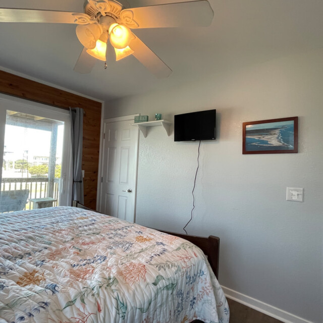 Bedroom with ceiling fan, floral bedspread, wall-mounted TV, framed art, and door to a bathroom. Balcony visible outside.