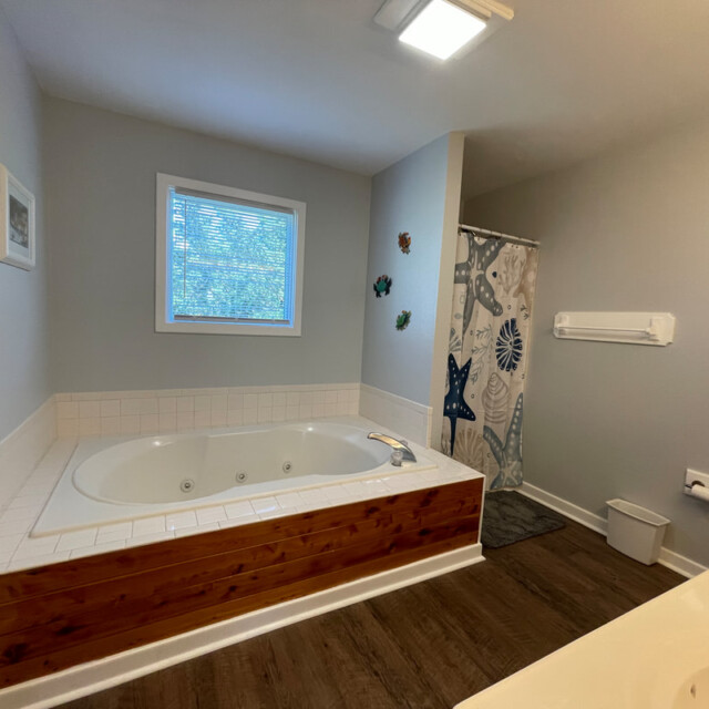 Bathroom with a large corner bathtub, window, shower with ocean-themed curtain, and wood-look flooring.