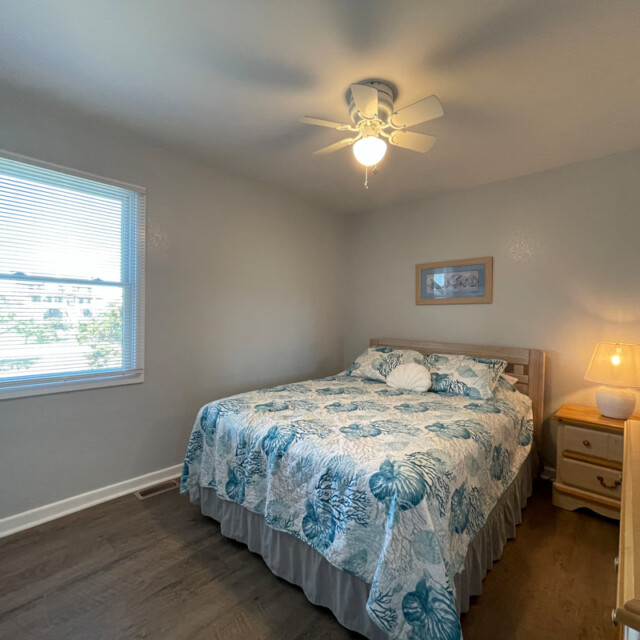 Small bedroom with a bed, floral bedding, nightstand with lamp, dresser, window, and ceiling fan.