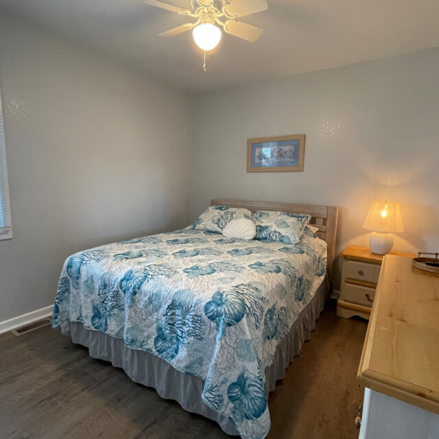 Bedroom with a queen bed, floral bedding, nightstand with lamp, dresser, window, and ceiling fan.