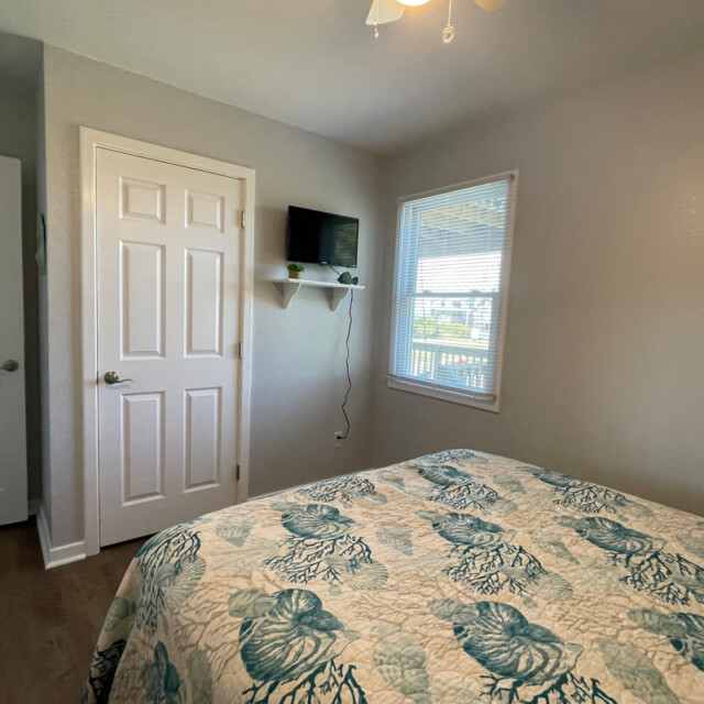 Small bedroom with a bed, patterned quilt, two closed doors, a wall-mounted TV, and a window with daylight.