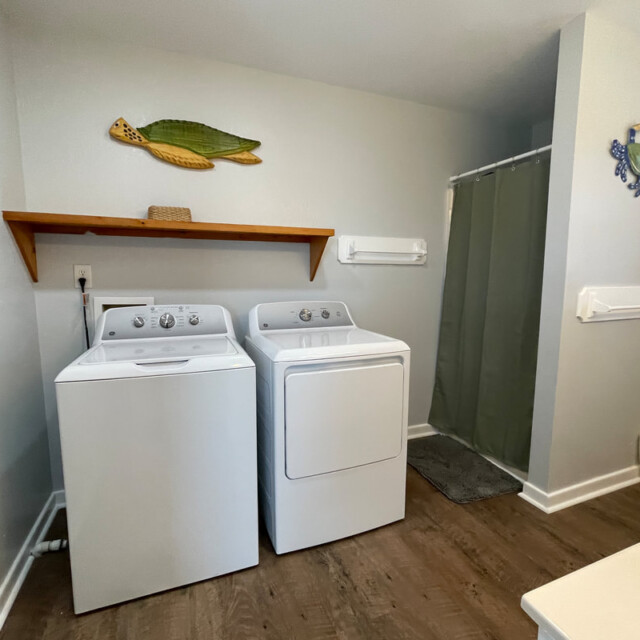 Laundry area with a washing machine and dryer, wooden shelf above, and a shower with a green curtain in the background.