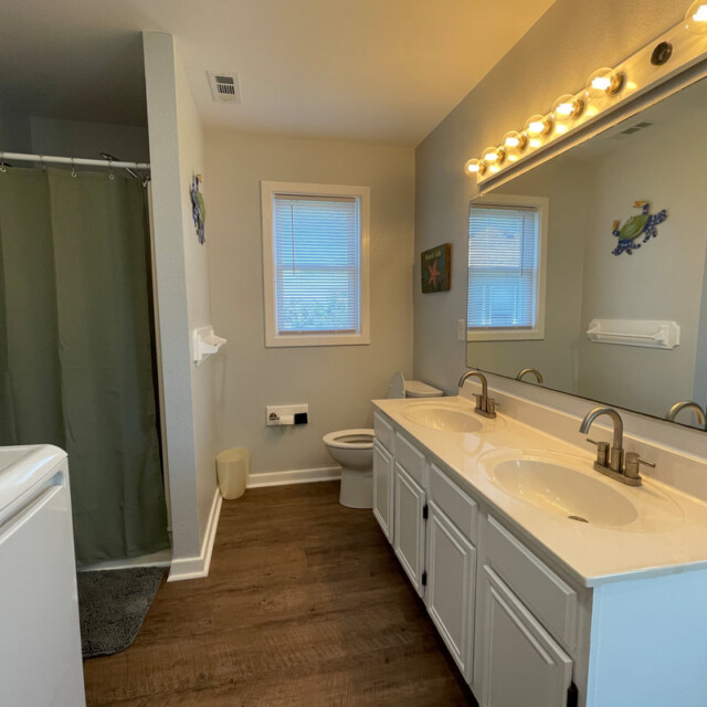 Bathroom with double sinks, a large mirror, a toilet, a shower with green curtain, and wood-style flooring.