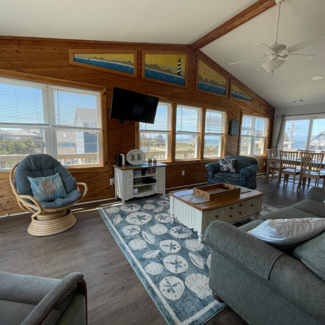 Bright living room with large windows, coastal decor, wood walls, a gray sofa, armchair, and dining area in the background.