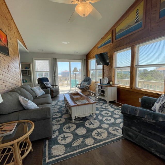 Living room with large windows, coastal decor, sofas, a coffee table, TV, and view of deck and houses outside.
