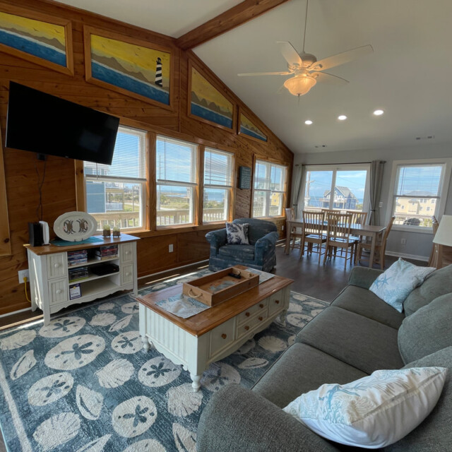 Living room with wood walls, large windows, gray sofa, TV, dining table, ceiling fan, and beach-themed decor.