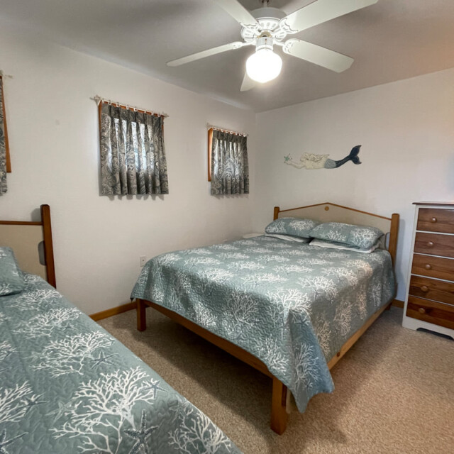 Bedroom with a double bed, a twin bed, wooden dresser, ceiling fan, and small windows with patterned curtains.