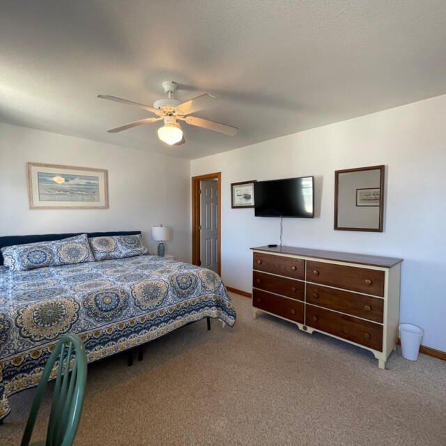 Bedroom with a patterned bedspread, ceiling fan, dresser, wall-mounted TV, and a window letting in natural light.