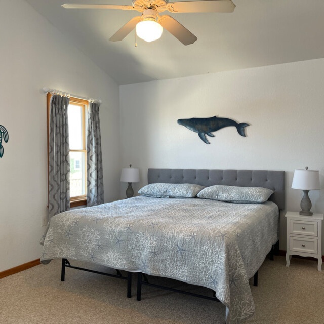 Bedroom with a king bed, two nightstands, sea-themed wall art, window with curtains, and a ceiling fan.
