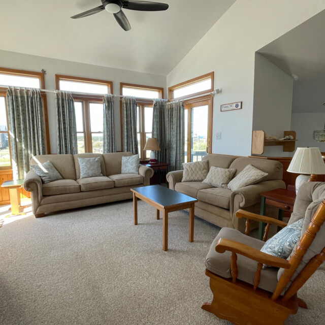 Bright living room with two beige sofas, a rocking chair, coffee table, and multiple large windows with curtains.