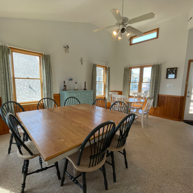 Bright dining area with a wooden table, eight chairs, sideboard, windows, and ceiling fan in a coastal-themed room.