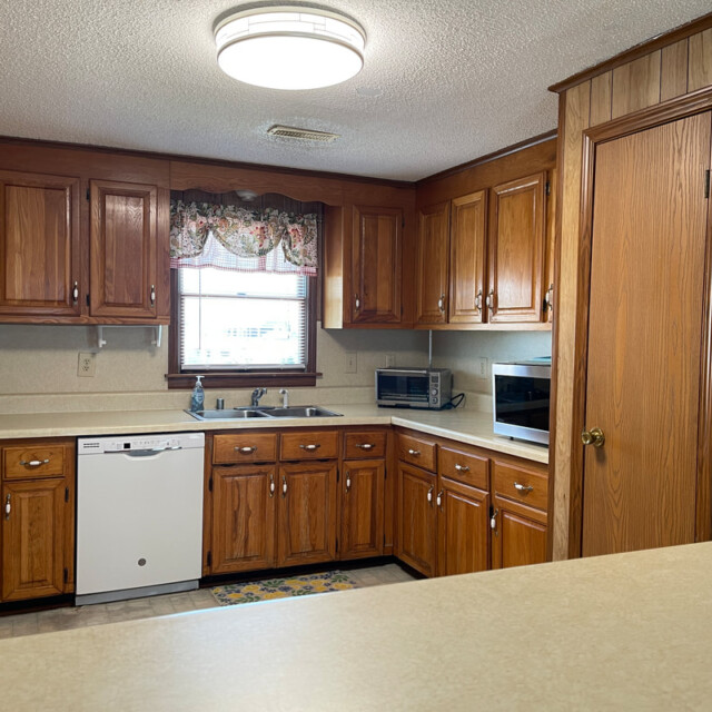 Kitchen with wood cabinets, a white dishwasher, microwave, and sink beneath a window with a floral valance.
