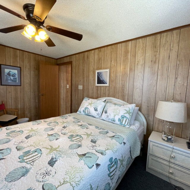 Bedroom with wood-paneled walls, a double bed with sea-themed bedding, nightstand, lamp, chair, and ceiling fan.