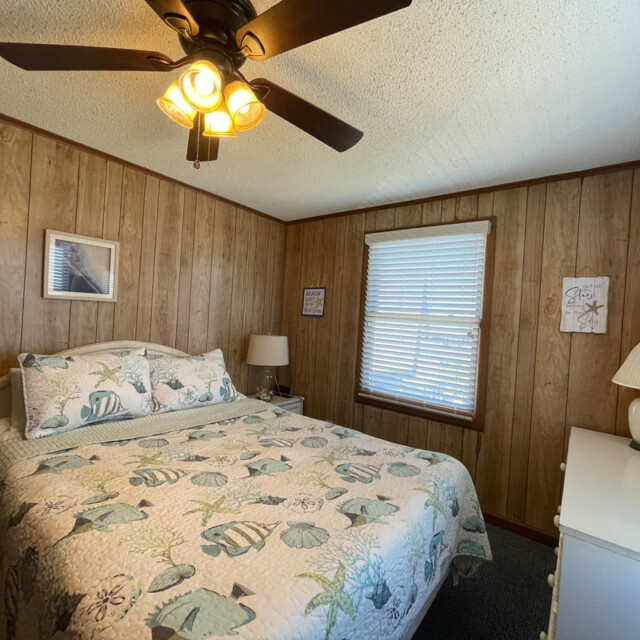 Bedroom with wood-paneled walls, a bed with a fish-themed quilt, a dresser, lamps, and a ceiling fan.