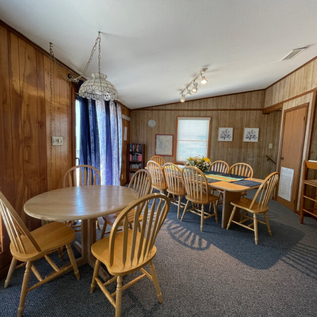 Dining area with two wooden tables, several chairs, wood-paneled walls, and windows with blinds and curtains.