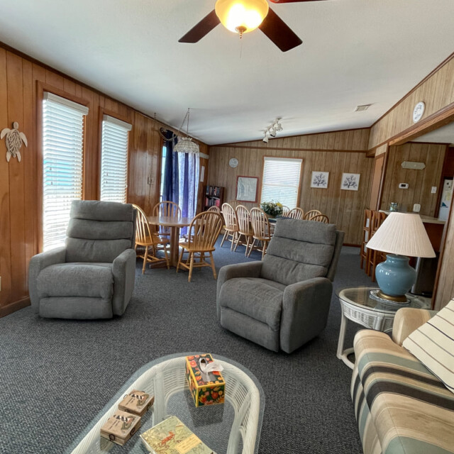 Living room with wood-paneled walls, two gray recliners, dining table, striped sofa, and ceiling fan.