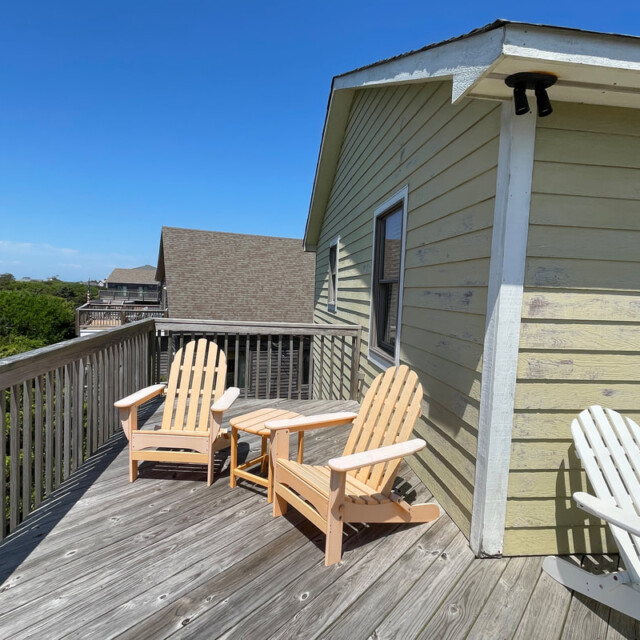 Wooden deck with three Adirondack chairs next to a yellow house, under a clear blue sky.