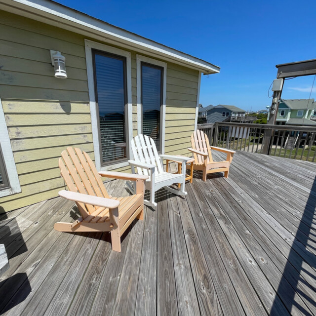 Three wooden chairs sit on a sunlit wooden deck outside a light green house, with neighborhood houses in the background.