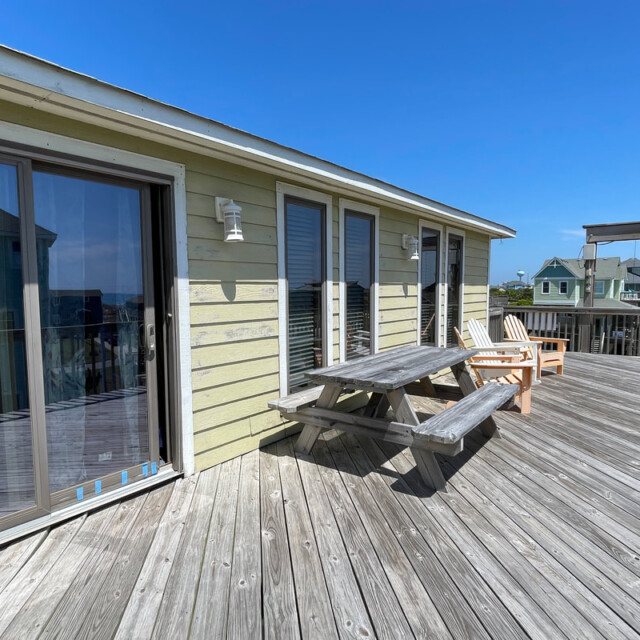Wooden deck with a picnic table and chairs outside a light yellow house, under a clear blue sky.