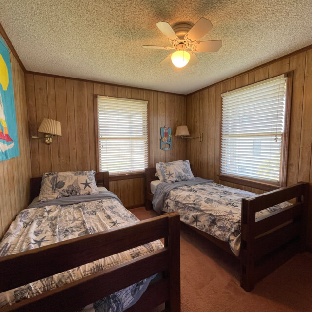 Small bedroom with wood-paneled walls, two twin beds, nautical decor, and light from two windows.